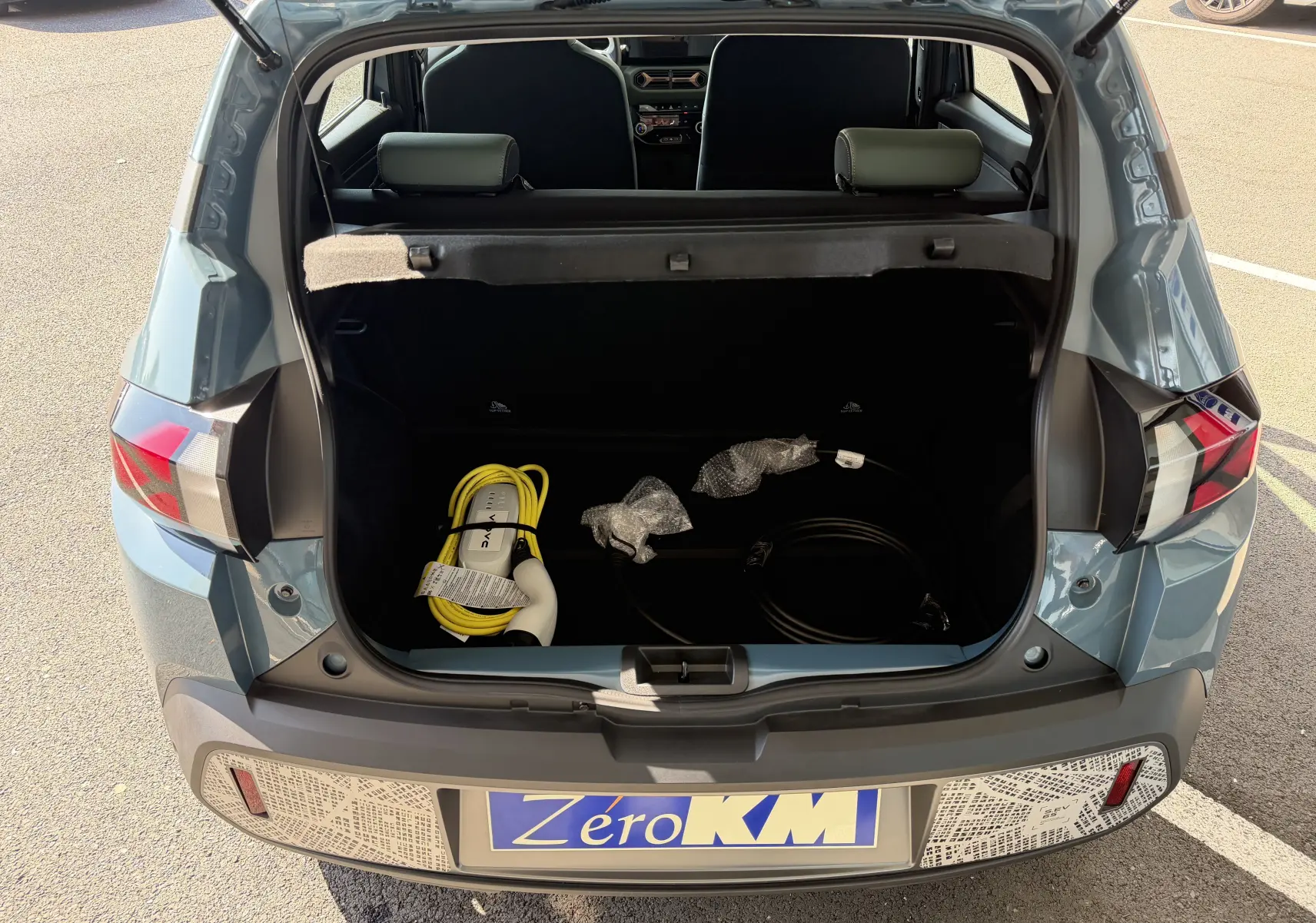 Coffre ouvert vu de l'arrière d'une Dacia Spring 65 Extreme bleu ardoise 2026 avec câbles de recharge visibles.
