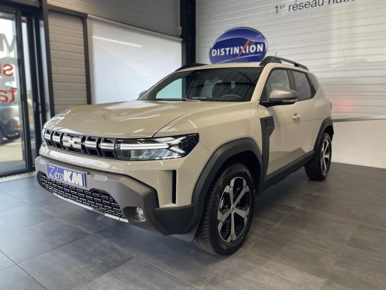 SUV Dacia Duster beige en 3/4 avant droit, avec jantes alliage noires et barres de toit noires visibles en showroom.