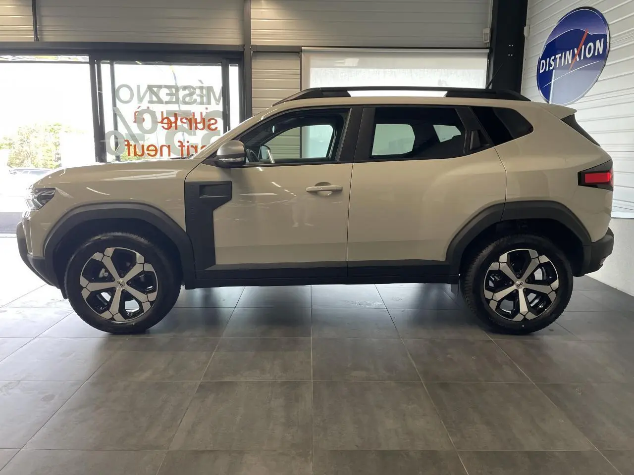 Profil côté gauche du SUV Dacia Duster beige Sandstone 2025 avec jantes alliage 18 pouces et barres de toit noires.