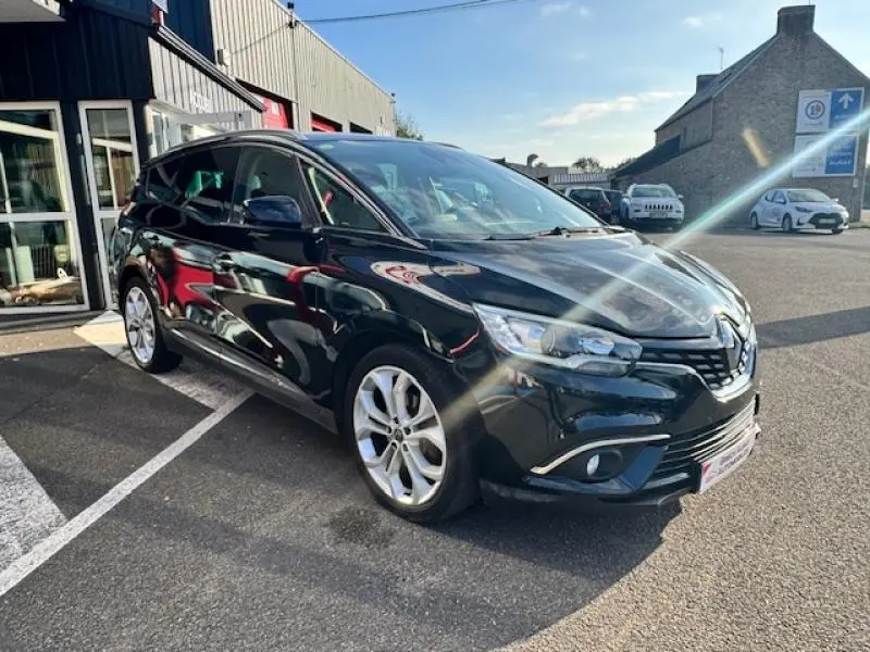 Renault Grand Scenic noir étoilé en 3/4 avant droit, stationné devant un bâtiment industriel par temps ensoleillé.