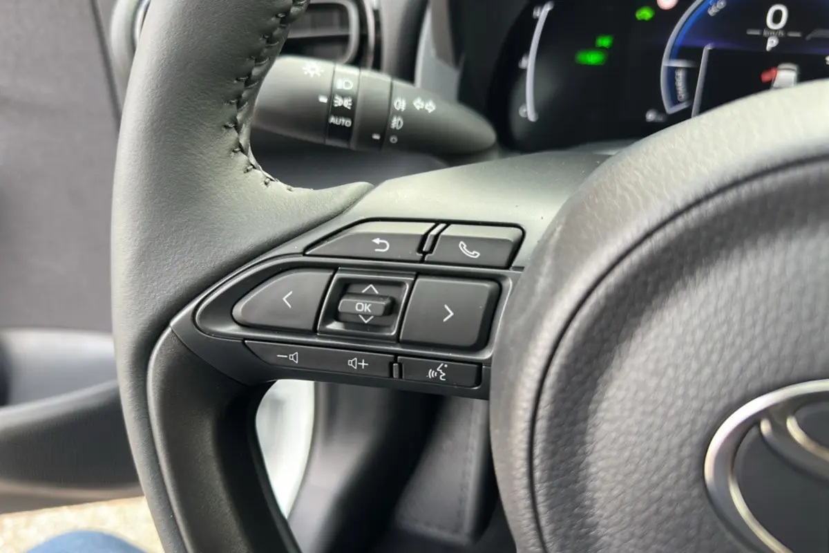 Gros plan sur les commandes du volant noir de la Toyota Yaris 1.5 Hybrid 116 BVA Design Plus 2025, tableau de bord en arrière-plan.
