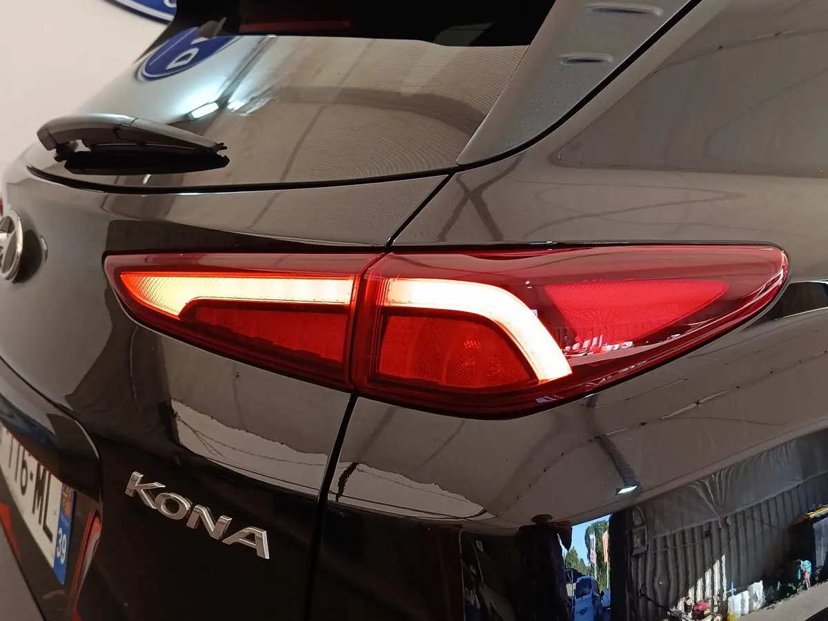 Gros plan sur le feu arrière droit allumé d'une Hyundai Kona électrique noire, avec logo et reflet d'atelier visible.