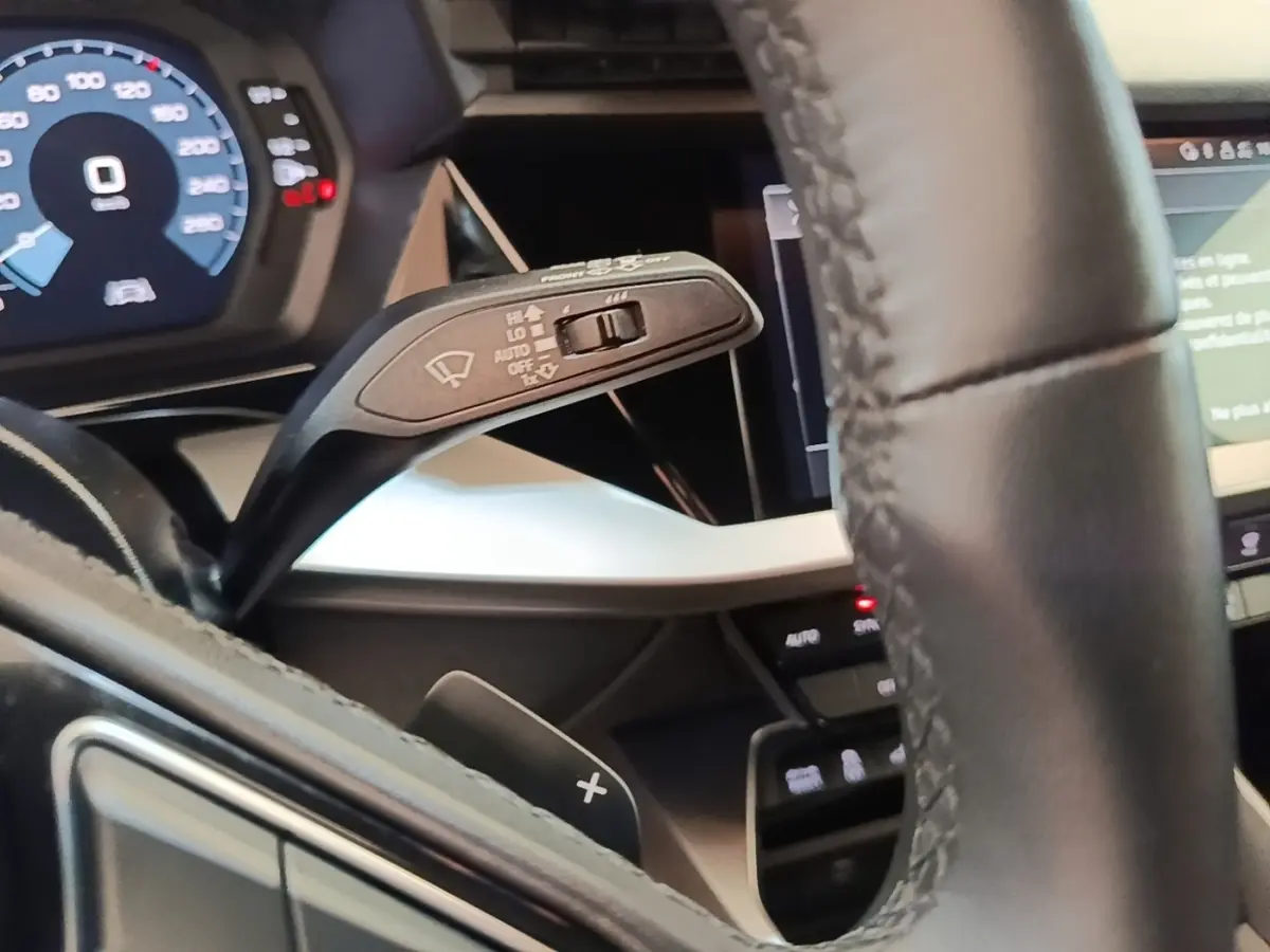 Gros plan sur le volant cuir noir et le commodo d’essuie-glace de l’Audi A3 hybride blanche, tableau de bord numérique visible.