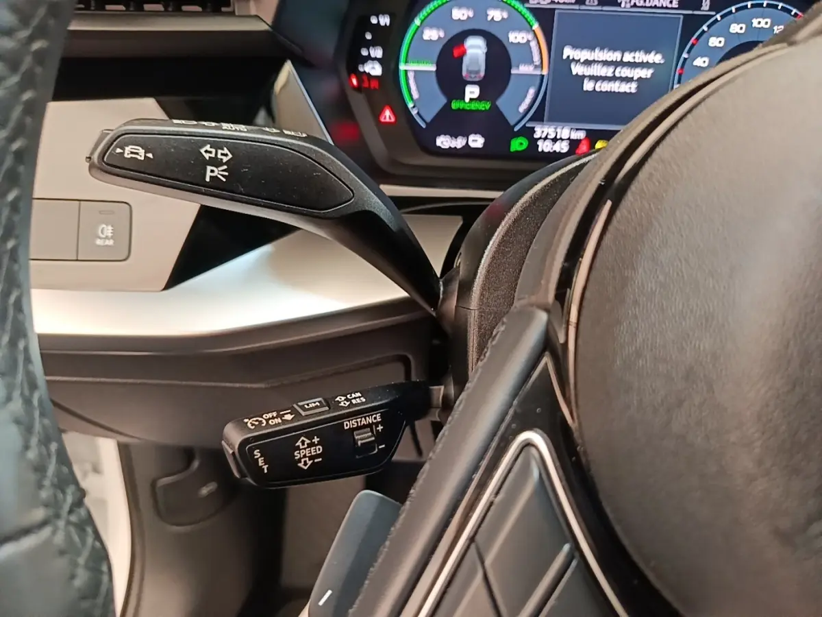 Gros plan sur le volant noir et les commandes au volant d'une Audi A3 blanche hybride rechargeable, tableau de bord digital visible.