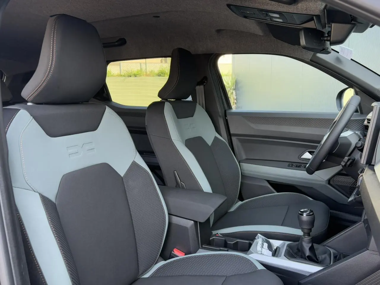 Vue intérieure côté conducteur du Dacia Duster 2025 avec sièges bi-ton gris clair et noir et levier de vitesse manuel.