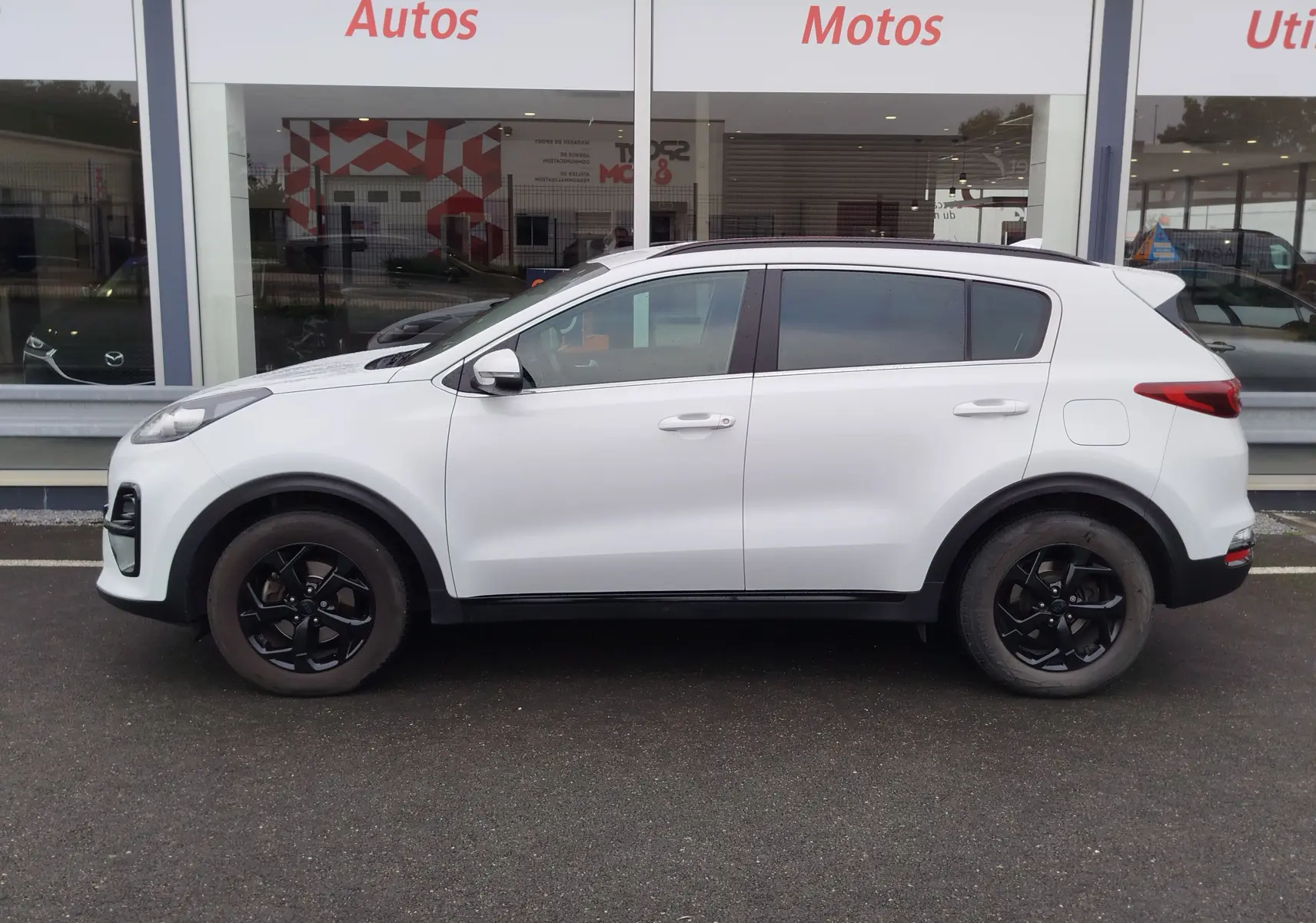 Profil côté gauche du KIA Sportage blanc 2022 avec jantes noires et vitres teintées, stationné devant un showroom.
