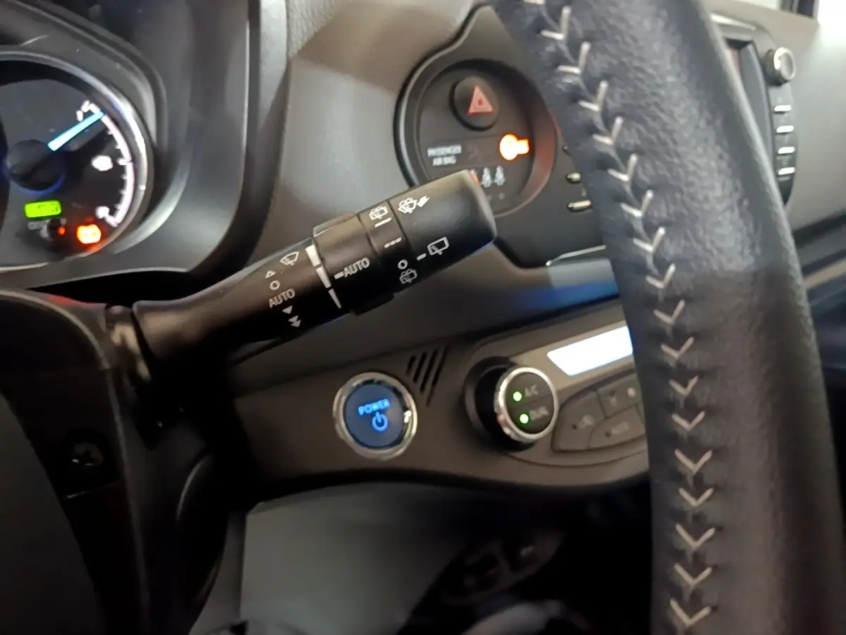Gros plan intérieur sur le volant cuir et la commande essuie-glaces de la Toyota Yaris hybride blanche, avec bouton démarrage bleu.