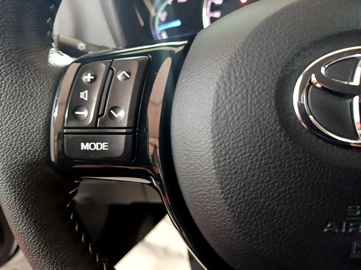 Gros plan sur les commandes gauche du volant en cuir noir de la Toyota Yaris hybride blanche, avec logo Toyota visible.