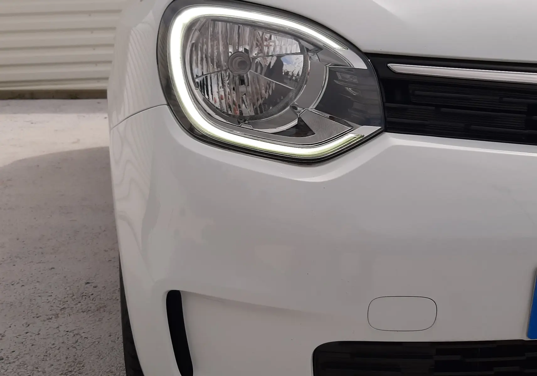 Gros plan sur le phare avant gauche allumé de la Renault Twingo III blanc cristal, avec calandre noire et carrosserie lisse.