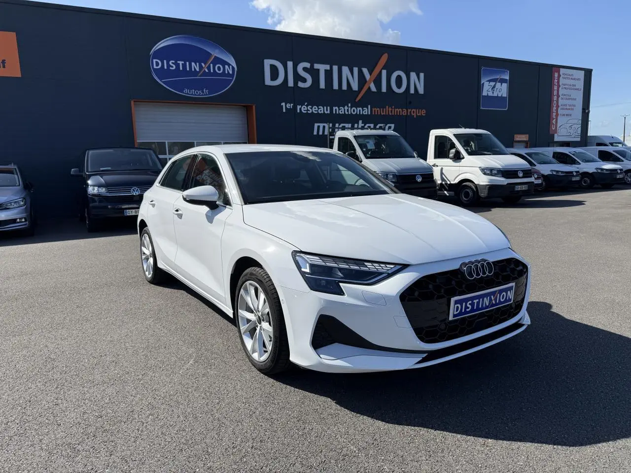 Audi A3 Sportback blanc Arcona vue 3/4 avant droit sur parking avec bâtiment Distinxion en arrière-plan.