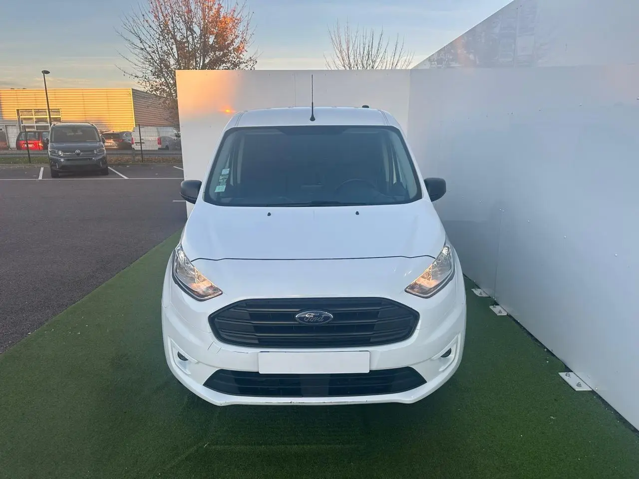 Vue frontale d'un fourgon Ford Transit Connect blanc avec calandre noire et phares allumés.