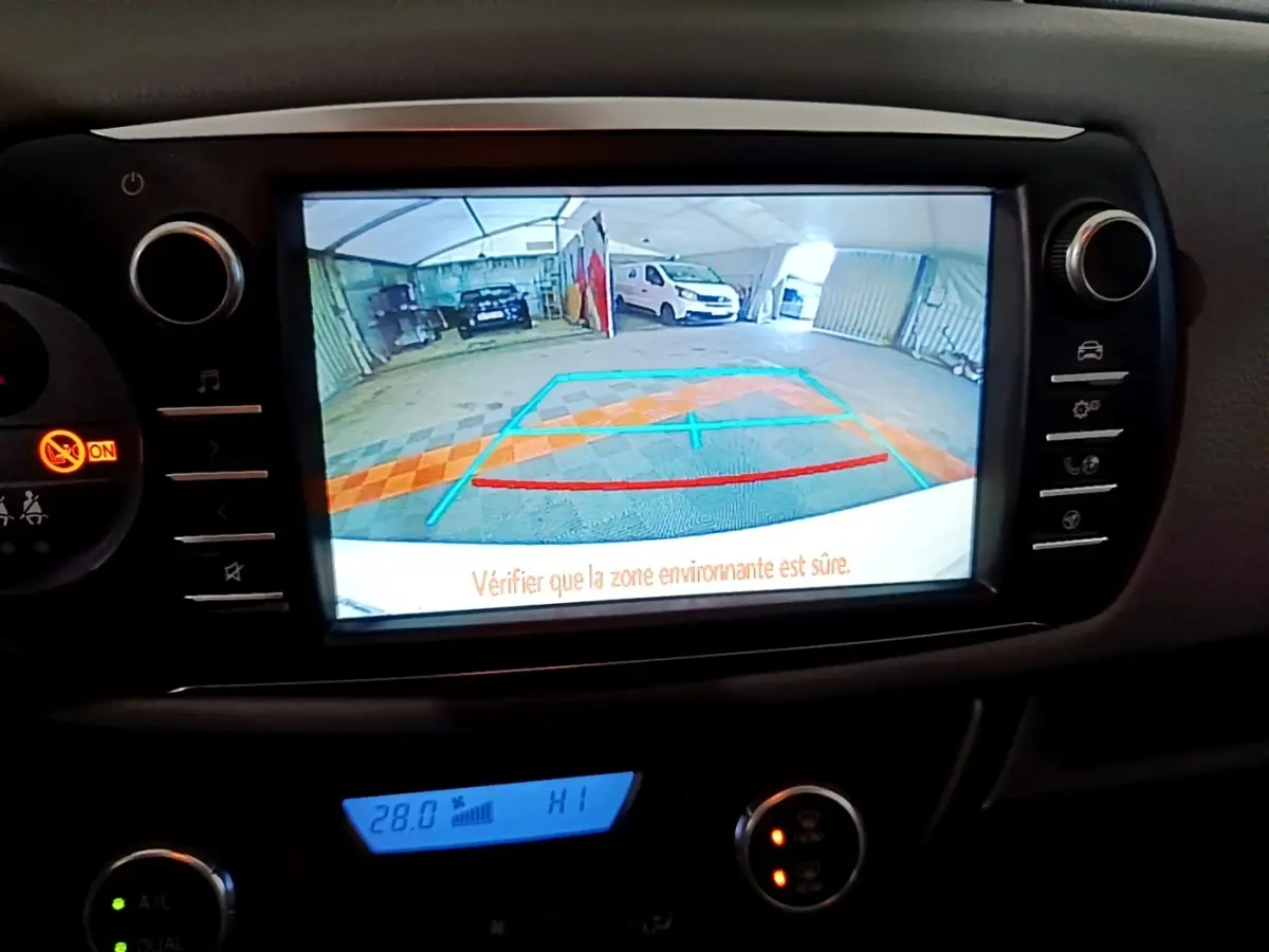 Écran tactile central affichant la caméra de recul d'une Toyota Yaris blanche, vue intérieure du tableau de bord.
