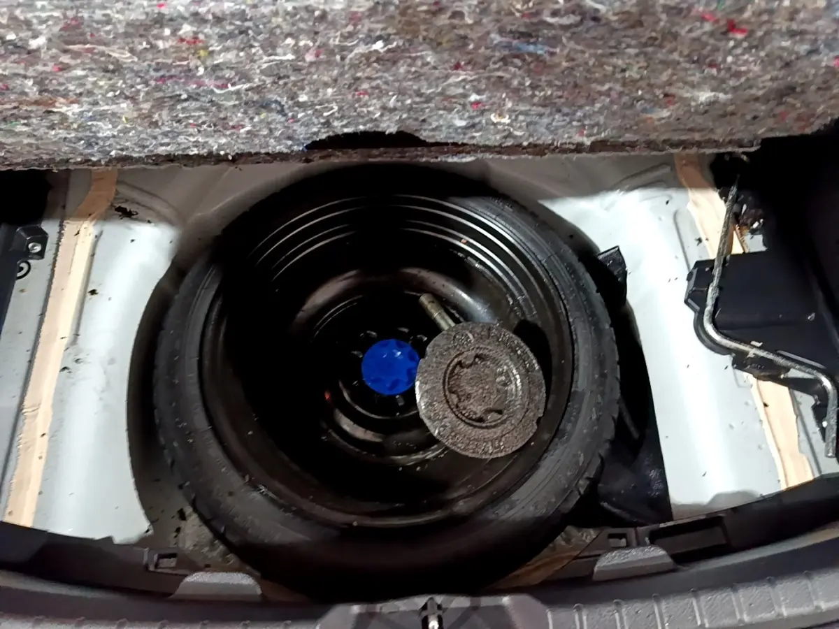 Vue plongeante sur la roue de secours noire dans le coffre d'une Toyota Yaris blanche, avec tapis de coffre partiellement visible.