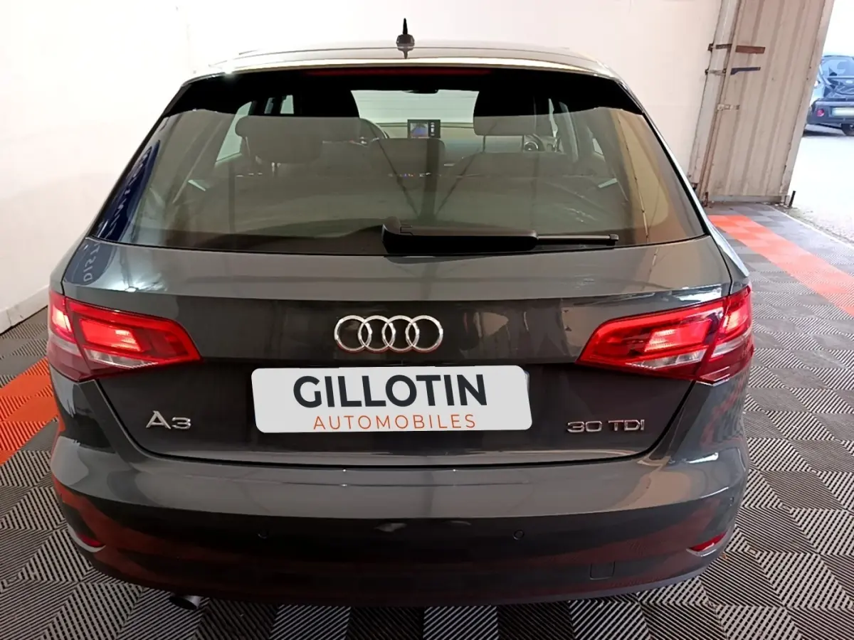 Vue arrière d'une Audi A3 30 TDI gris foncé, feux arrière allumés, dans un garage avec sol noir et rouge.