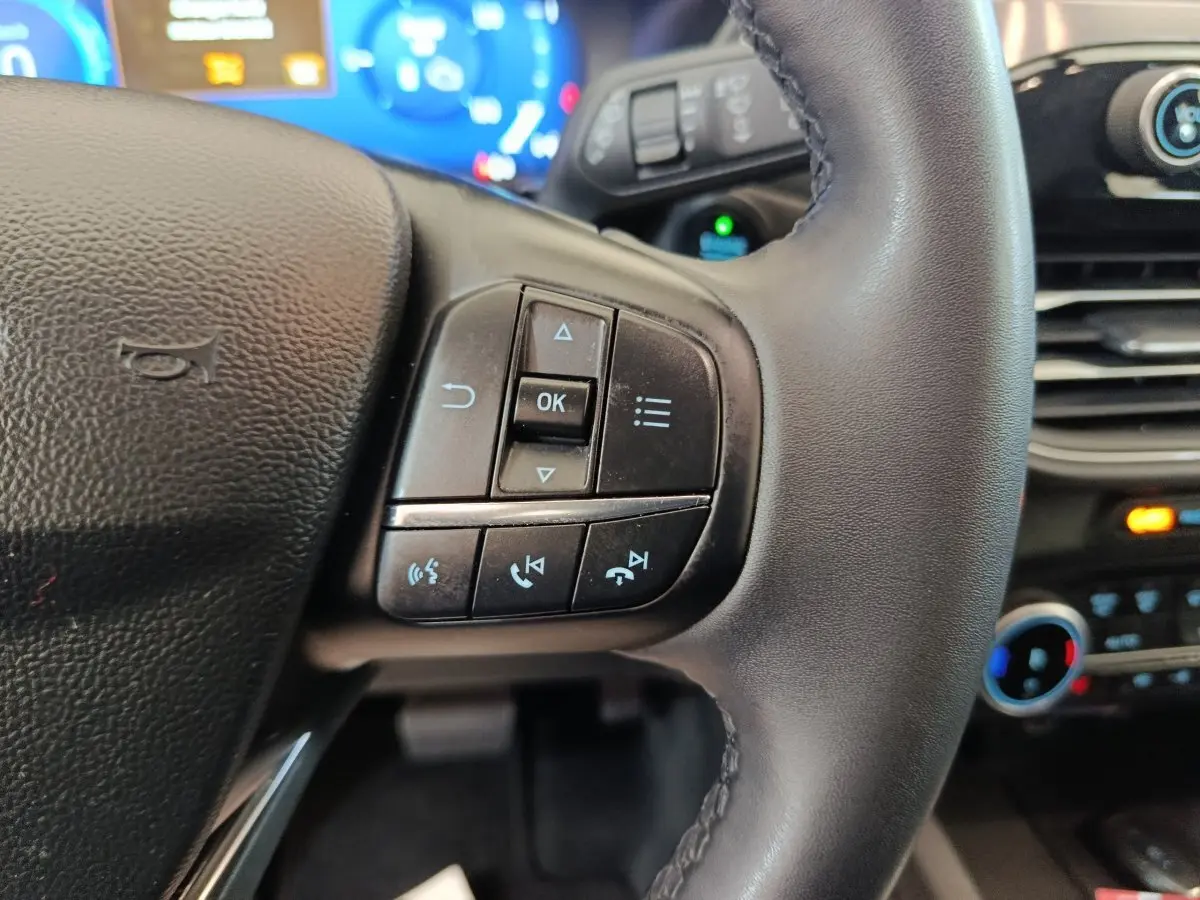 Gros plan sur les commandes gauche du volant cuir noir du Ford Kuga 2023, avec tableau de bord numérique bleu en arrière-plan.