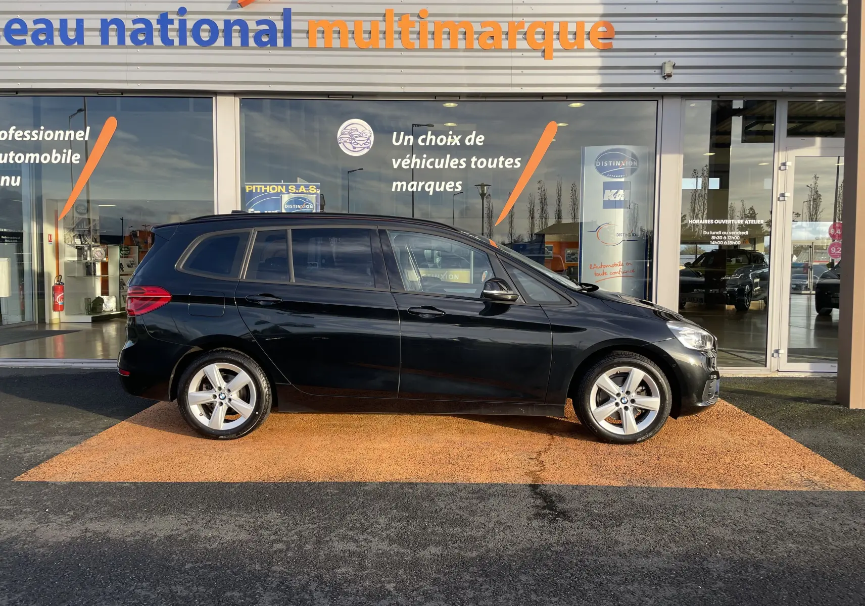 Profil côté gauche d'une BMW Série 2 Gran Tourer F46 noire, garée devant une concession multimarque en plein jour.