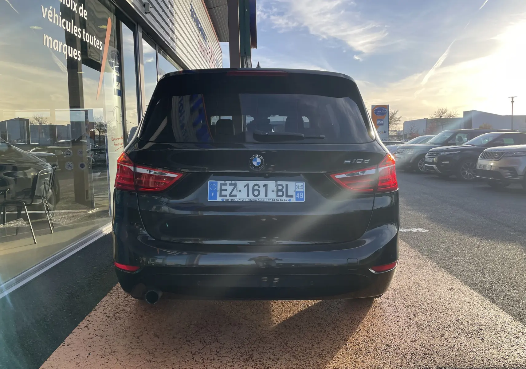 Vue arrière d'une BMW Série 2 Gran Tourer 218d noire, stationnée devant une vitrine en fin d'après-midi.