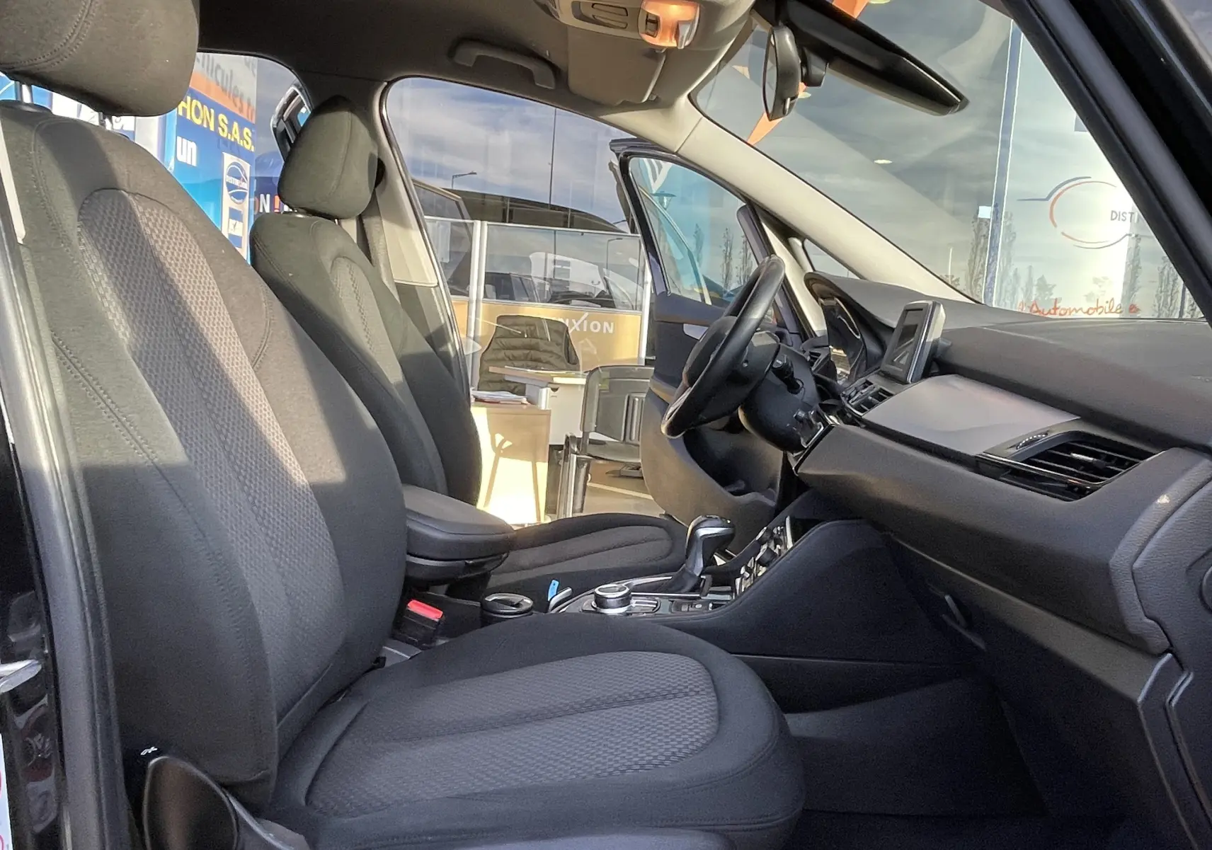 Vue intérieure côté conducteur de la BMW Série 2 Gran Tourer 218 DA 150 Lounge noire, montrant sièges et tableau de bord moderne.