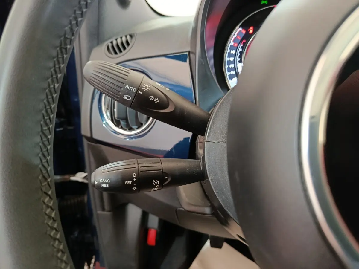 Détail intérieur côté gauche du volant cuir d’une FIAT 500 Dolcevita bleu, avec commandes de phares et régulateur.