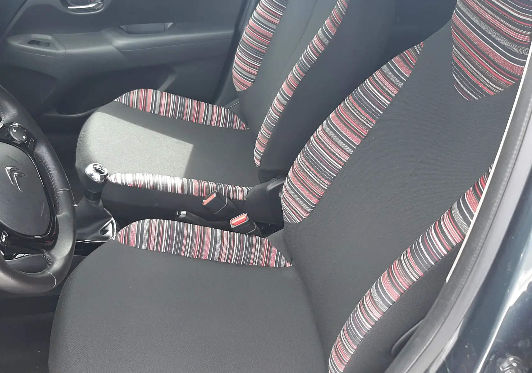 Vue intérieure côté conducteur de la Citroën C1 gris foncé 2019, sièges tissu noir avec bandes rayées rouges et blanches.