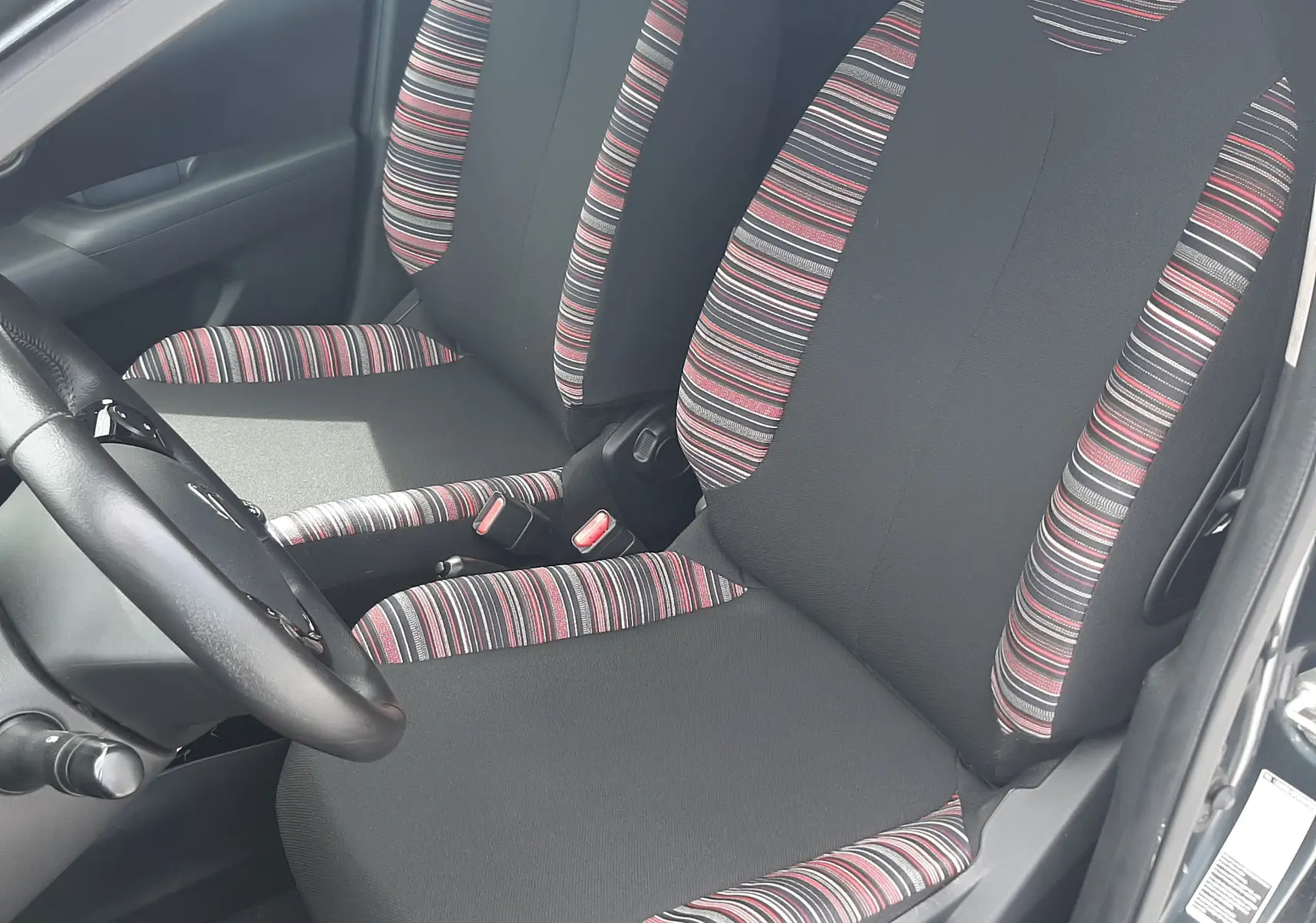 Vue intérieure côté conducteur de la Citroën C1 gris foncé 2019, sièges en tissu noir avec motifs rayés rouges et blancs.
