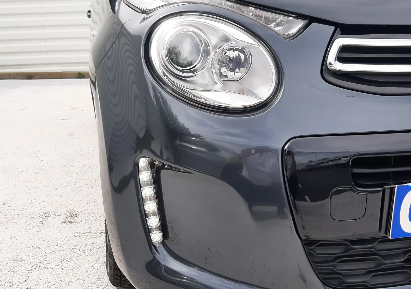 Gros plan sur l’avant gauche d’une Citroën C1 gris foncé 2019, mettant en valeur le phare rond et la bande LED verticale.