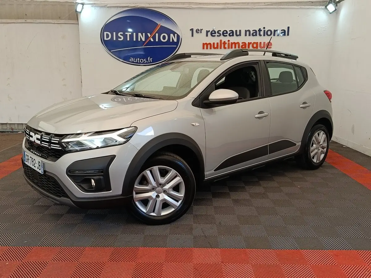 Vue 3/4 avant droit d'une Dacia Sandero Expression gris clair avec barres de toit et jantes alliage en showroom.