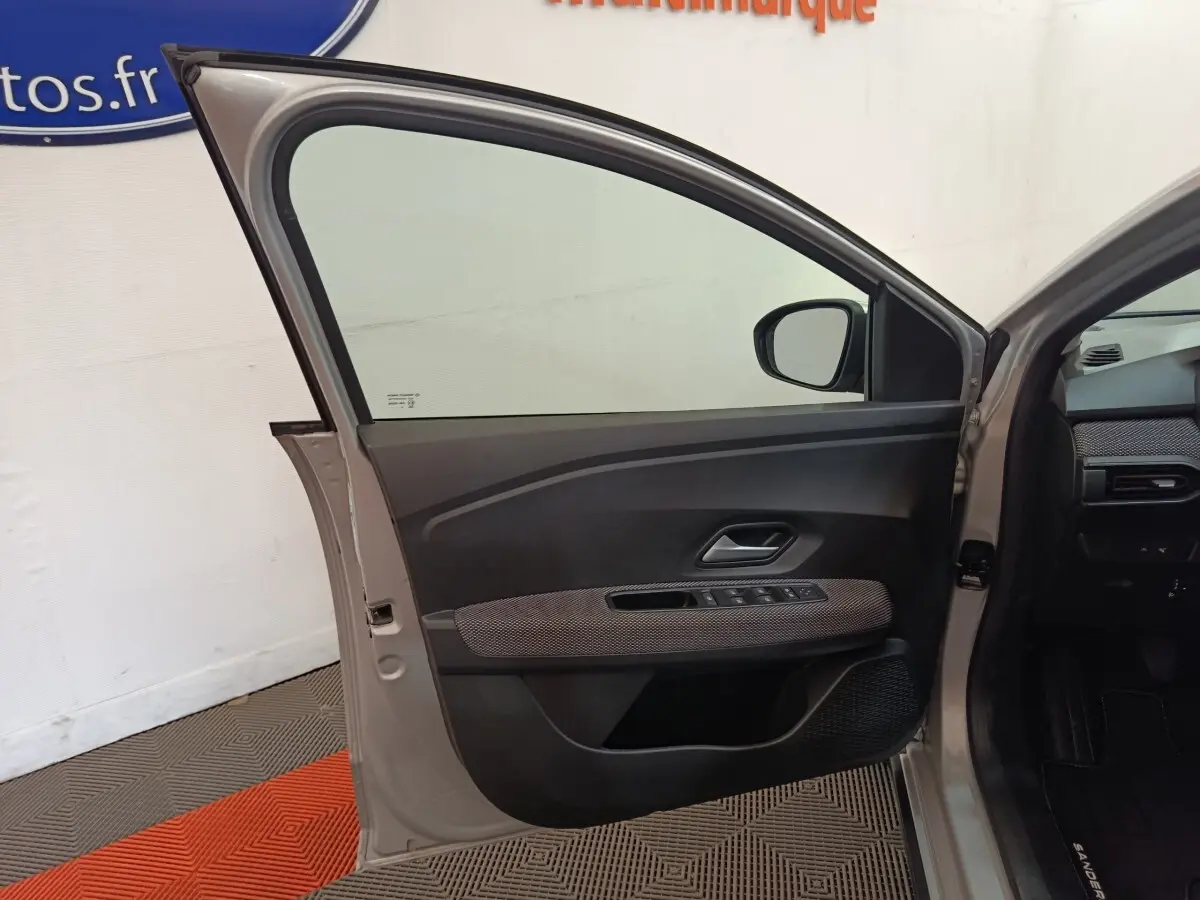 Porte avant gauche ouverte d'une Dacia Sandero gris clair 2023, montrant le panneau intérieur avec commandes électriques.