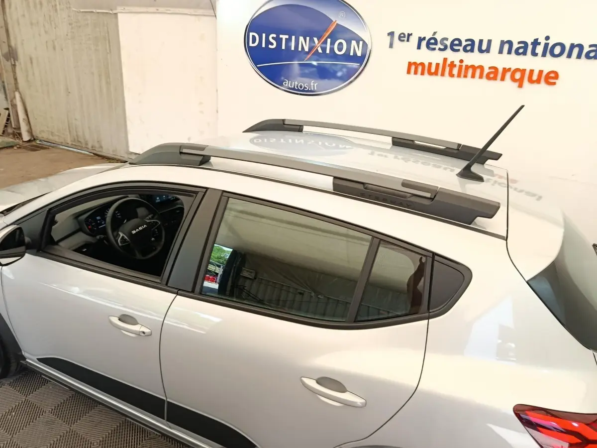 Vue latérale gauche d'une Dacia Sandero gris clair 2023 avec barres de toit et intérieur visible au volant multifonctions.
