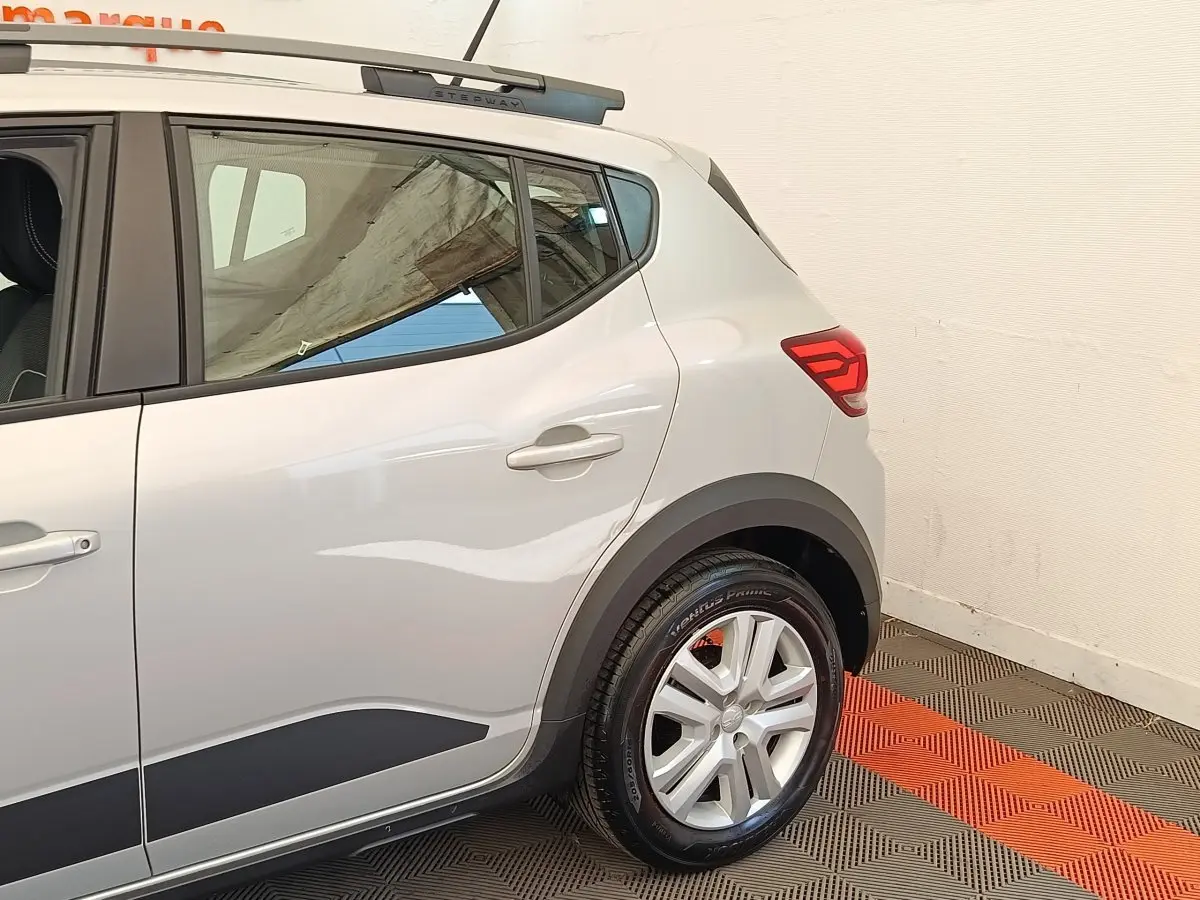Vue côté droit arrière d'une Dacia Sandero Expression gris clair avec jante alliage et barres de toit Stepway.