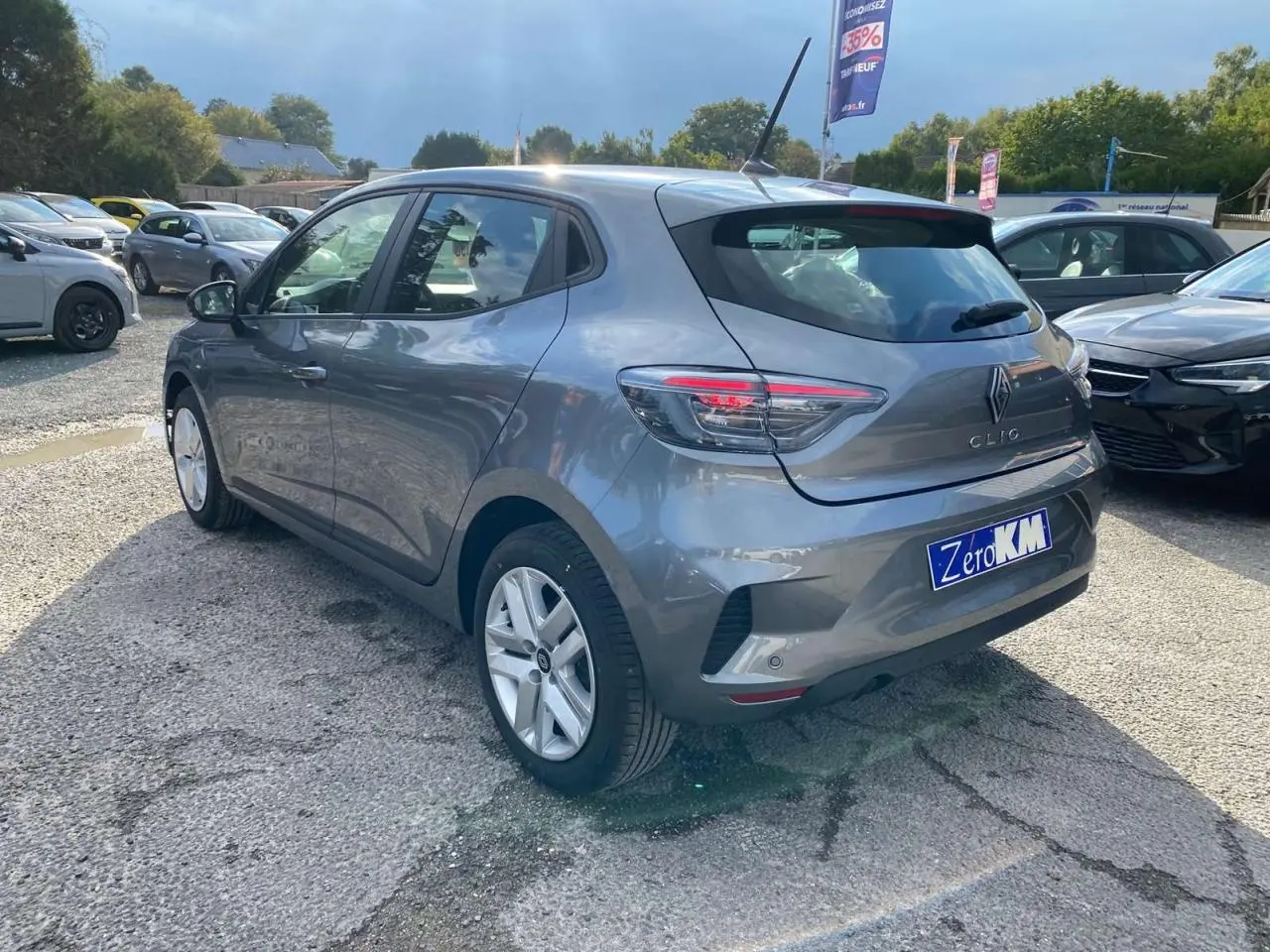 Renault Clio 2025 gris schiste vue 3/4 arrière droit sur parking avec feux LED et plaque Zero KM.