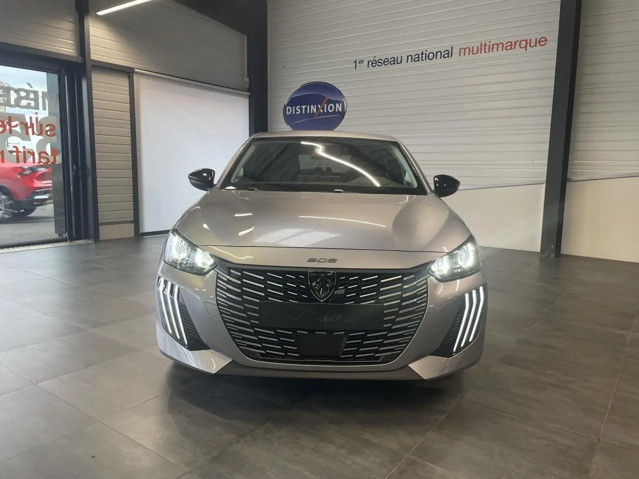 Vue frontale d'une Peugeot 208 1.2i Hybrid 2025 gris Artense avec phares LED allumés et calandre noire distinctive.