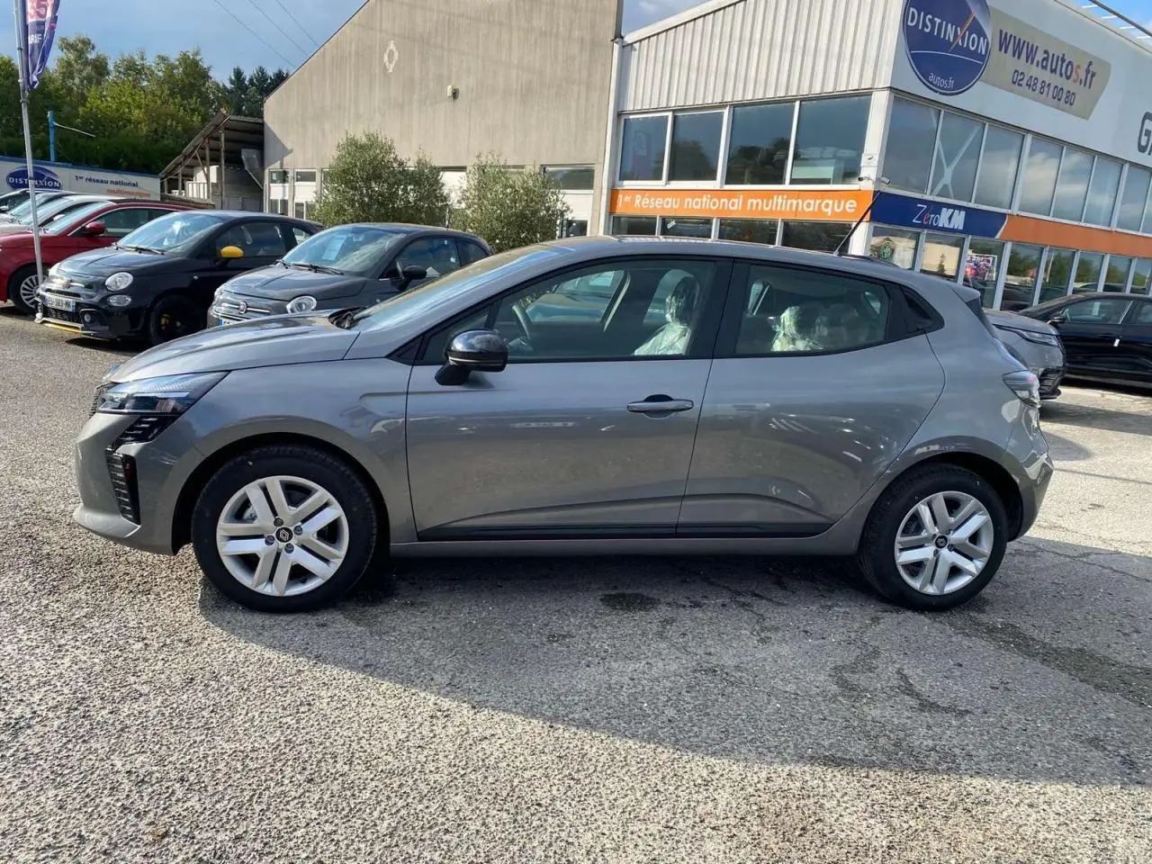 Profil droit d'une Renault Clio 2025 gris schiste garée devant un bâtiment commercial, avec jantes alliage visibles.