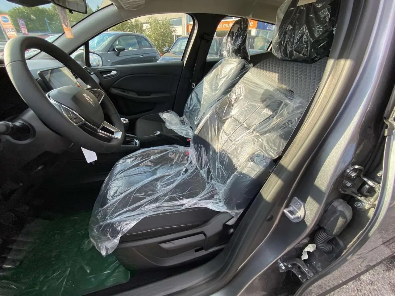 Intérieur côté conducteur de la Renault Clio gris schiste 2025, sièges avant protégés par film plastique, volant visible.