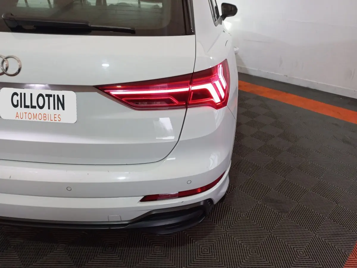 Vue arrière droite d'un Audi Q3 blanc 45 TFSI E S Line avec feux arrière LED allumés dans un garage.