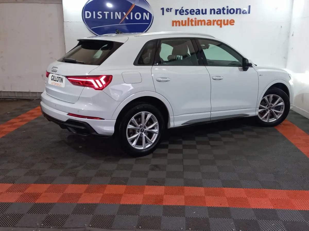 Audi Q3 blanc en vue 3/4 arrière droit, avec feux LED allumés et jantes alliage visibles en intérieur showroom.