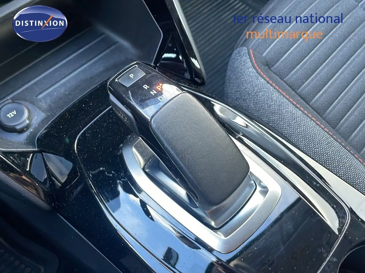 Le levier de vitesse automatique noir brillant de la Peugeot 208 électrique, avec siège tissu gris et surpiqûres rouges visible en arrière-plan.