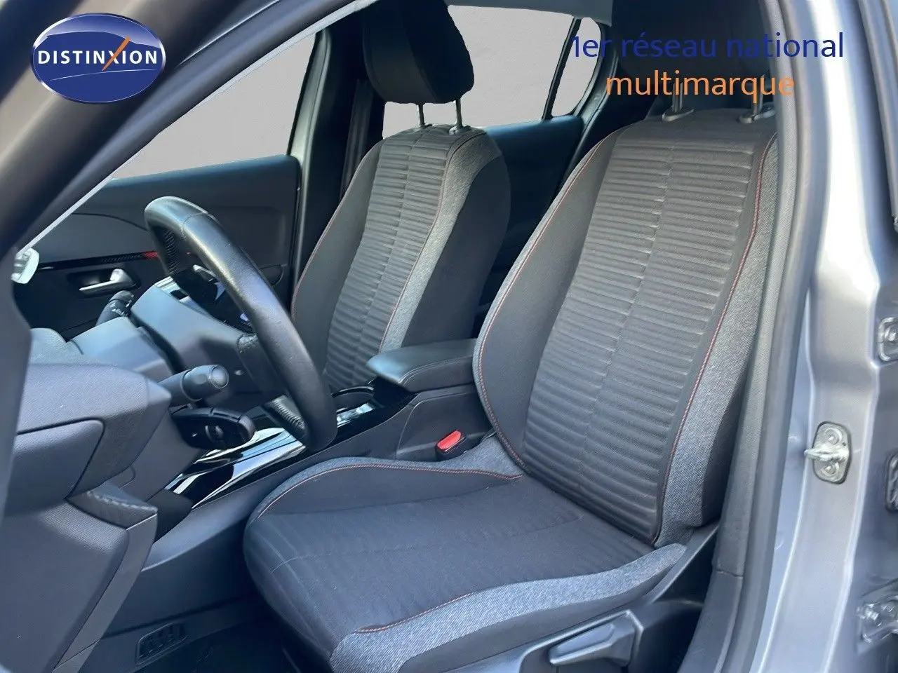 Vue intérieure côté conducteur de la Peugeot 208 électrique gris Artense, sièges tissu gris avec surpiqûres rouges.