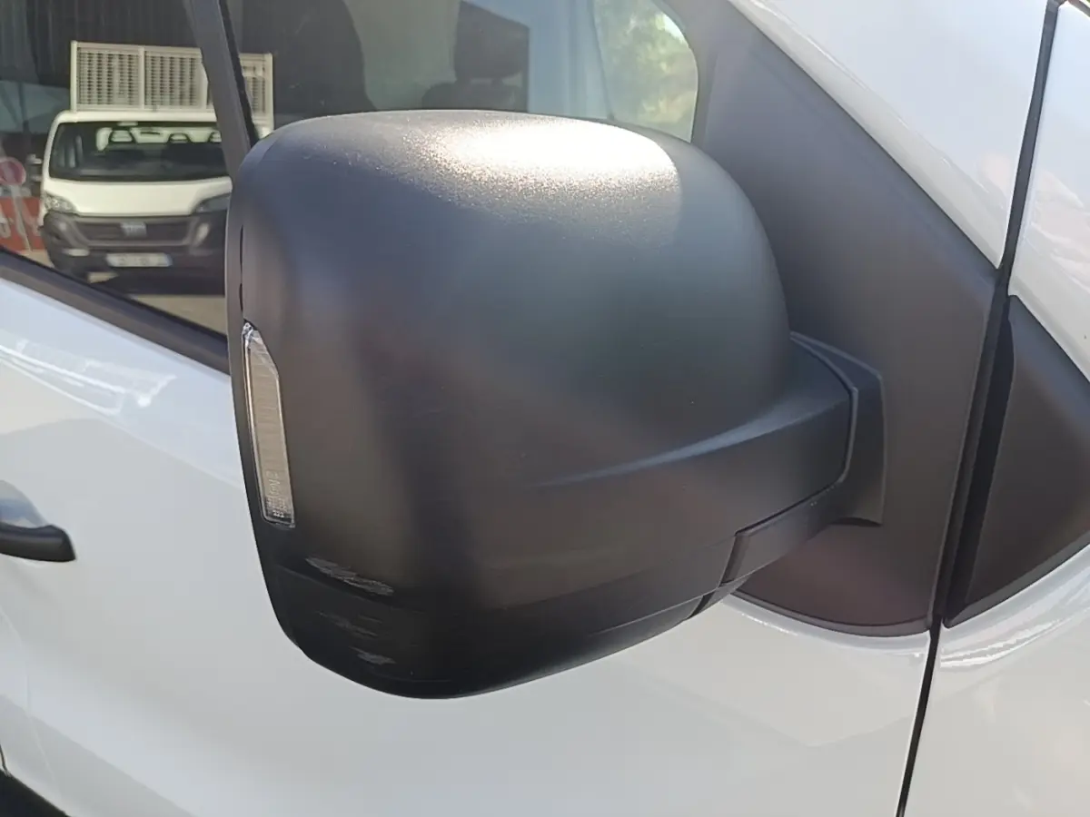 Gros plan sur le rétroviseur noir côté conducteur du Renault Trafic Fourgon blanc, avec reflet d’un autre véhicule en arrière-plan.