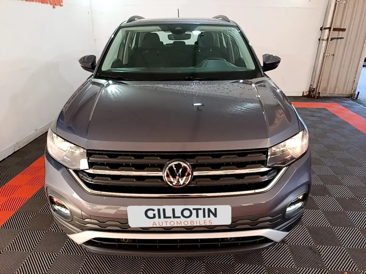 Vue avant d'un Volkswagen T-Cross gris foncé 2023 avec calandre chromée et feux allumés en intérieur.