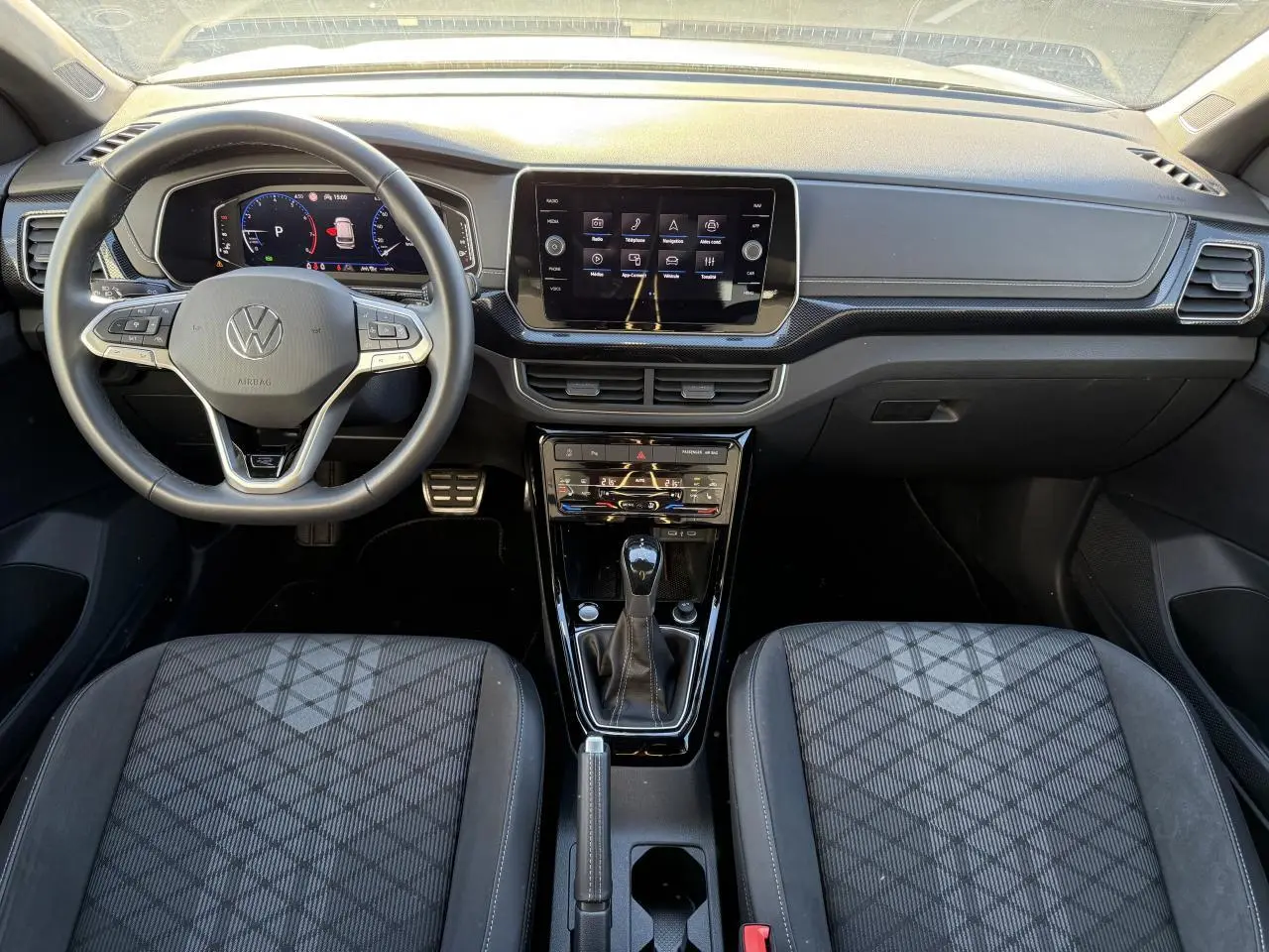 Vue intérieure avant du Volkswagen T-Cross 2025, tableau de bord noir avec écran tactile et sièges tissu à motifs géométriques.