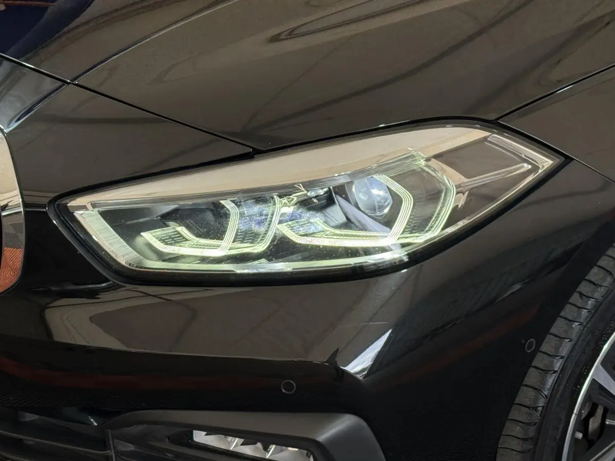 Gros plan sur le phare avant droit allumé d’une BMW Série 1 116d noire, avec partie de la roue visible.