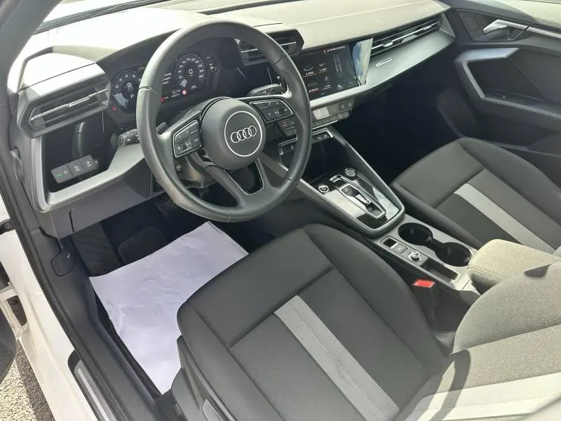 Intérieur avant de l'Audi A3 Sportback blanc Arkona 2024, volant cuir multifonction et sièges tissu noir avec bande grise.