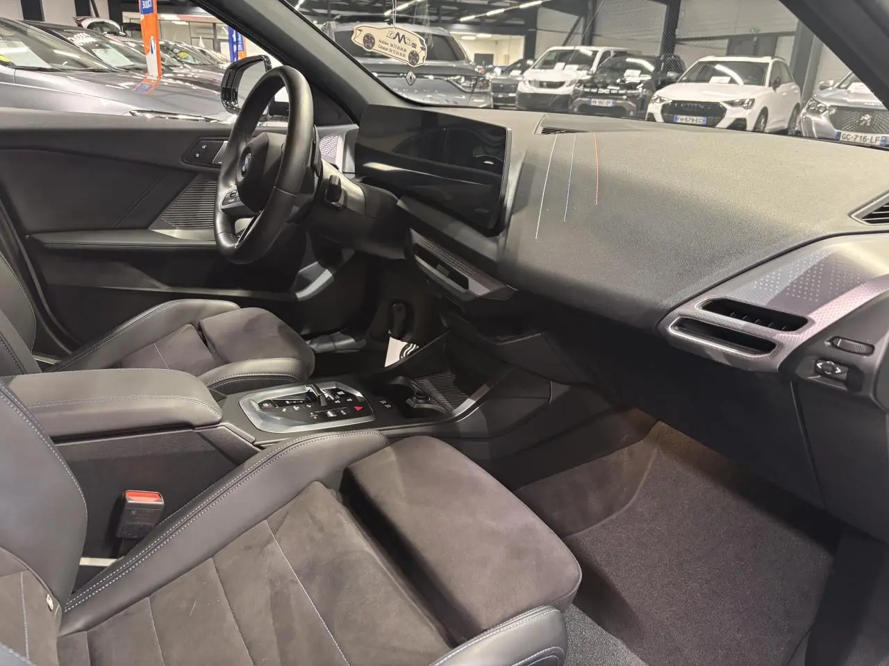 Intérieur BMW Série 1 118d 2025 vu côté conducteur, sellerie Alcantara noire avec surpiqûres bleues et tableau de bord moderne.