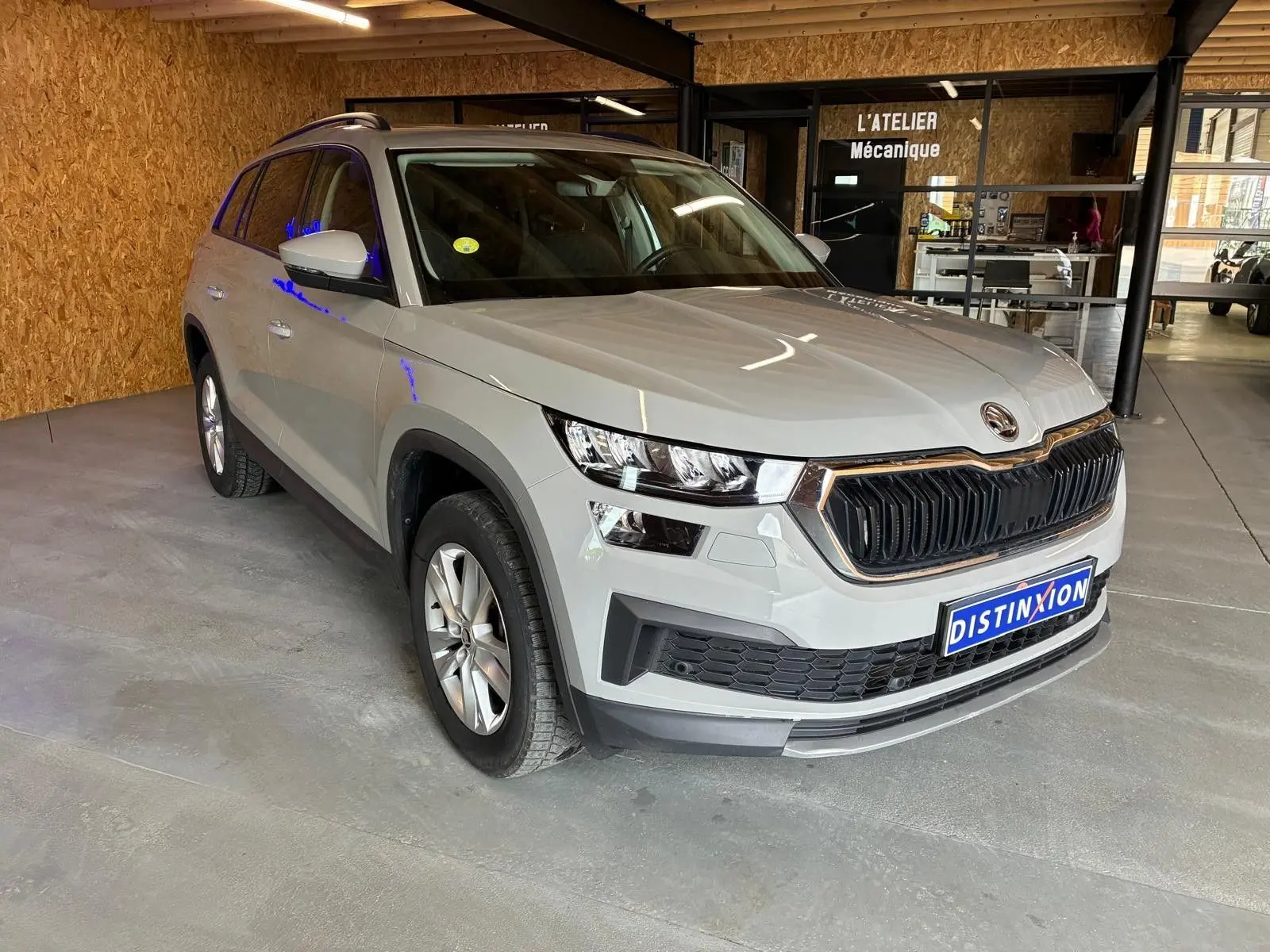SKODA KODIAQ gris clair en 3/4 avant droit, SUV 7 places avec calandre noire et jantes argentées, stationné en intérieur.