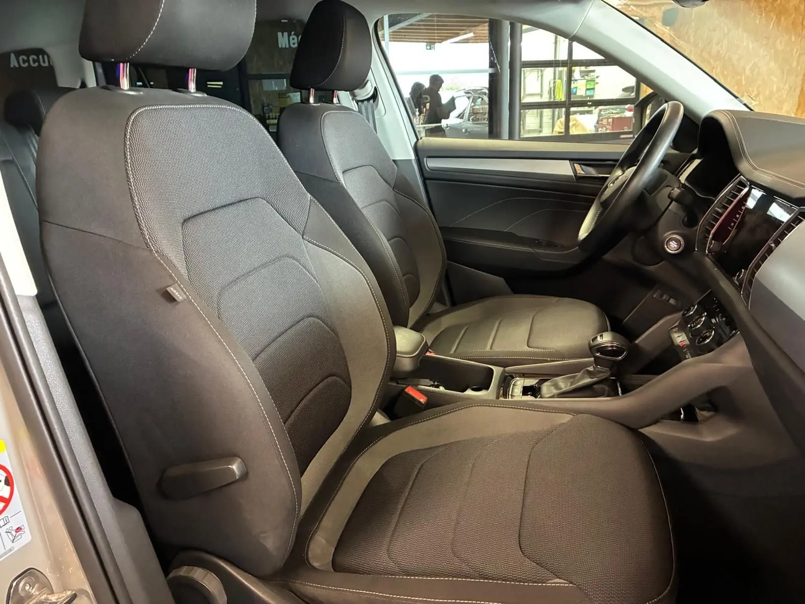 Vue intérieure côté gauche du Skoda Kodiaq 2023 gris clair, montrant les sièges avant en tissu noir et le tableau de bord.