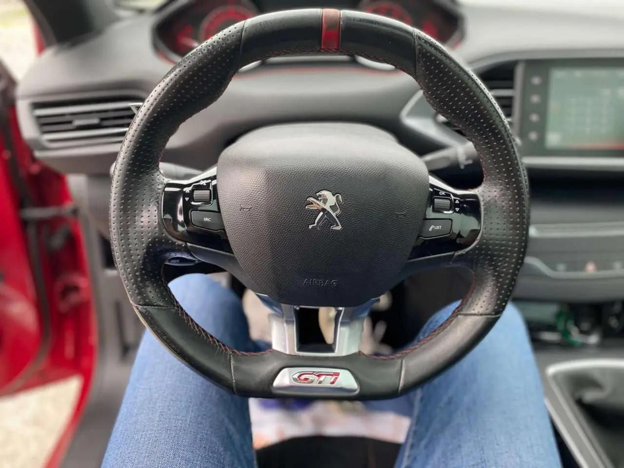 Volant cuir perforé avec logo GTi au centre d'une Peugeot 308 rouge, vue intérieure depuis le siège conducteur.