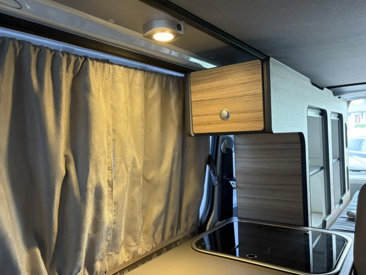 Intérieur du Volkswagen Transporter aménagé, vue latérale droite sur la kitchenette bois clair avec rideau beige fermé.