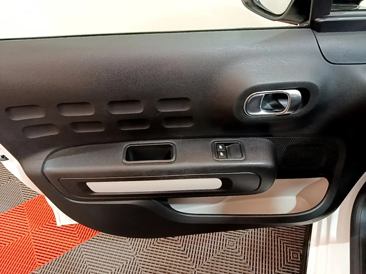 Intérieur de la porte avant gauche d'une Citroën C3 blanche, avec poignée argentée et commandes de vitres électriques.