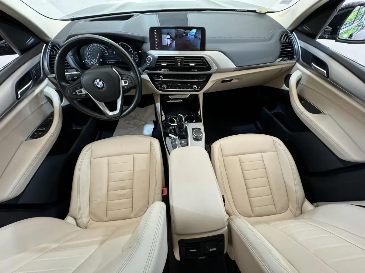 Vue intérieure avant de la BMW X3 2018 avec sièges cuir beige, tableau de bord noir et écran tactile central allumé.